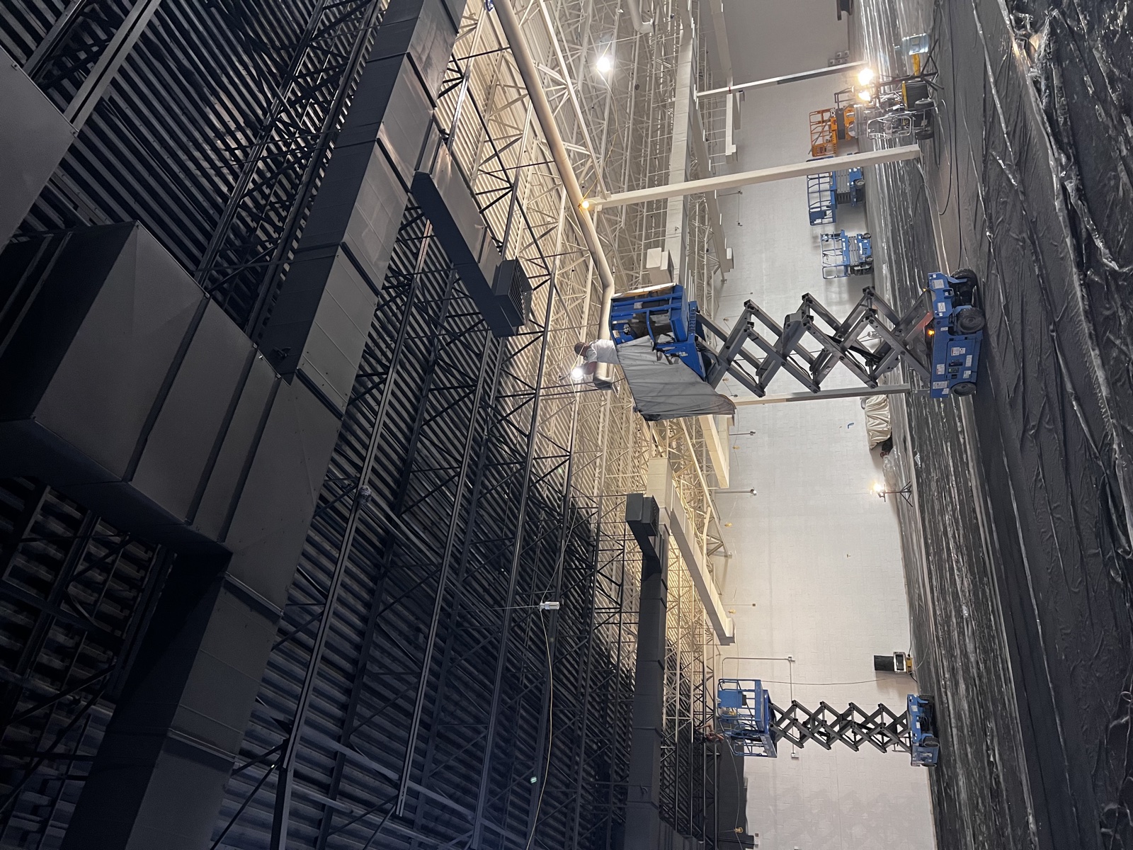 Large industrial distribution center interior with tall rebar shelving racks, gray polished concrete floor, and a blue scissor lift positioned for ceiling work