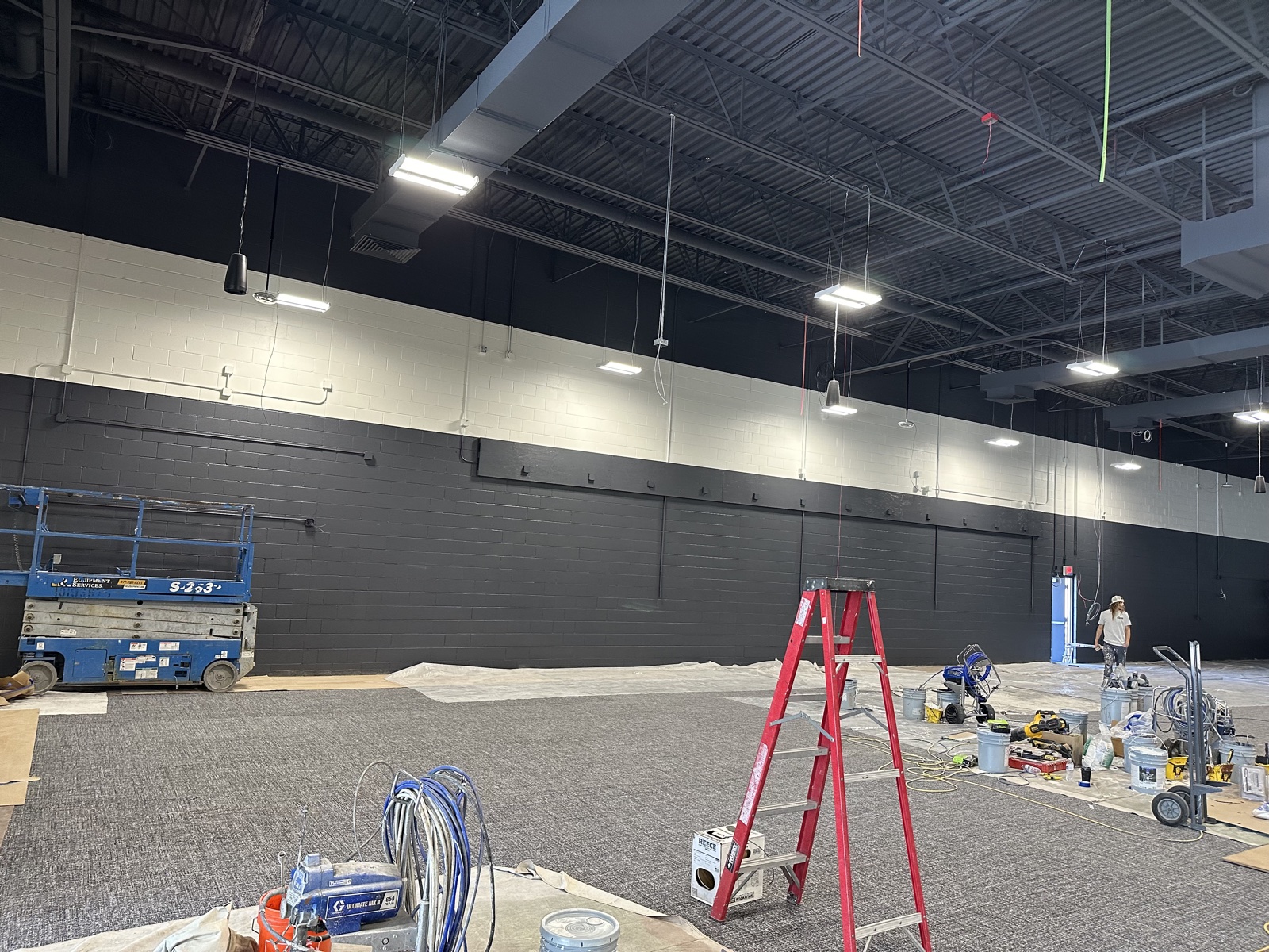 Large commercial space mid-repaint with a two-tone black-and-white wall scheme, drop cloths covering carpeted floor, and ladders and paint equipment in place