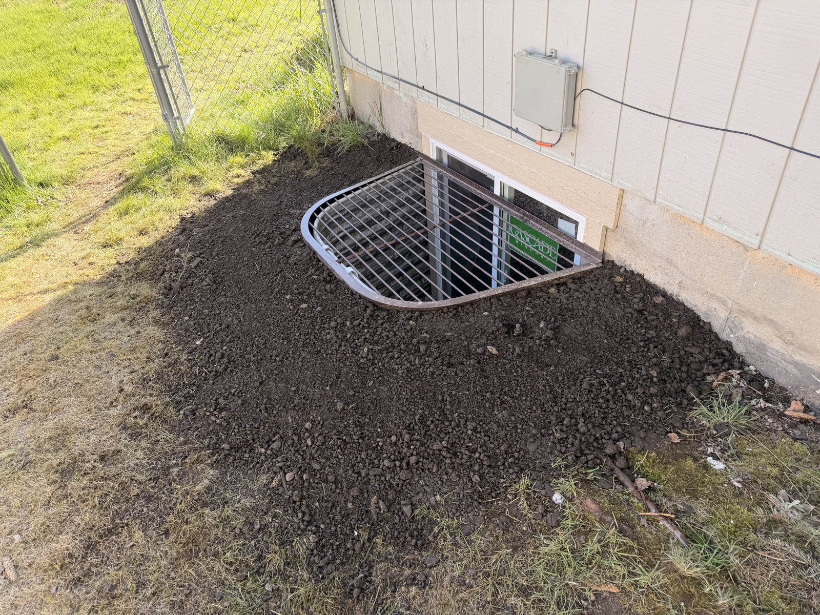 Galvanized metal egress window well newly bolted into a foundation cut on a tan-sided home, with fresh dirt mounded around the perimeter