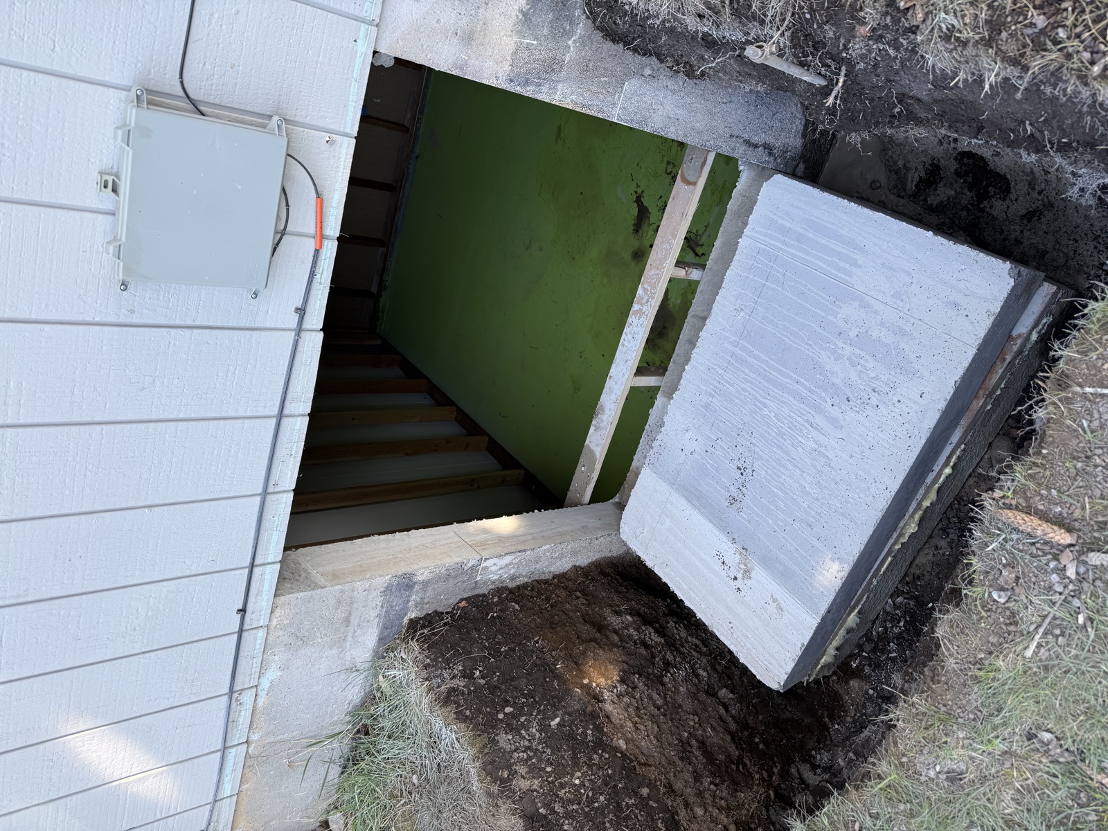 Completed metal egress window well at the base of a residential foundation with a concrete cap stone, wooden steps inside, and an exterior electrical panel adjacent