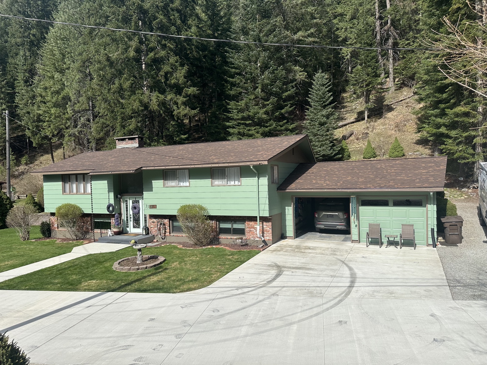 Sage green single-story ranch home with brown shingle roof, attached two-car garage, paved drive, and a forested mountain backdrop of pines