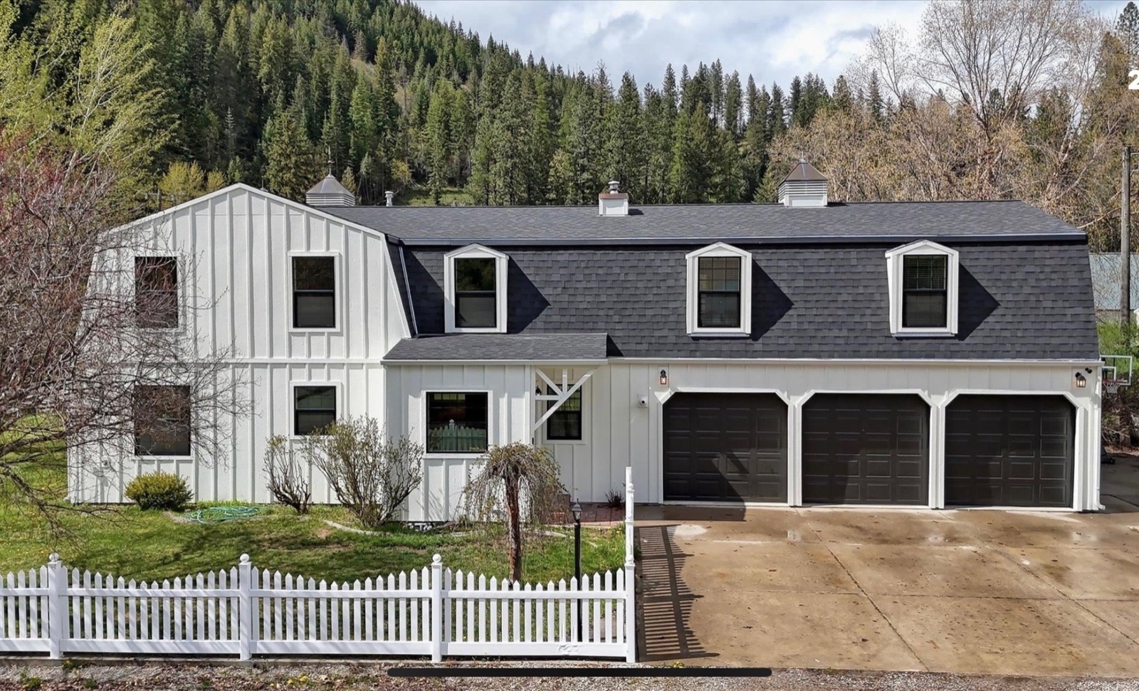 Two-story white modern farmhouse with gambrel-style roof, dormers, three-car black garage, and a white picket fence, set against a forested mountainside