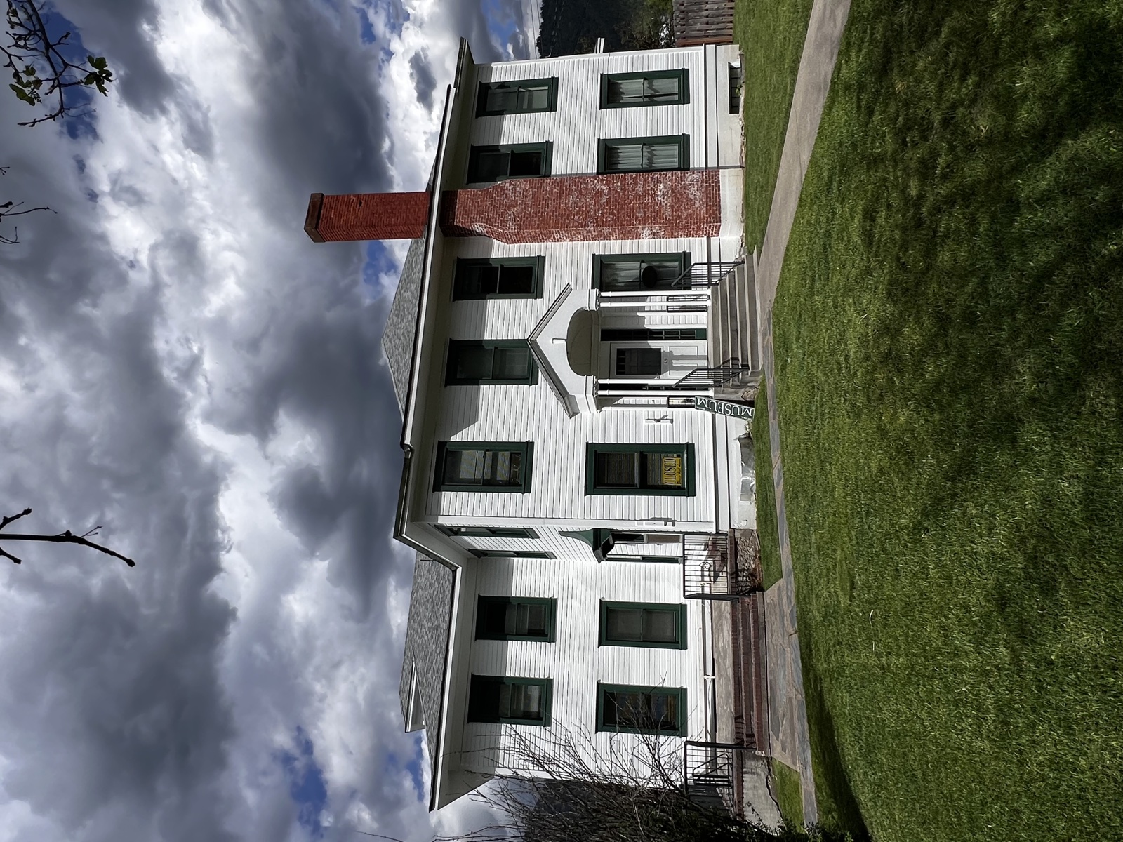 Two-story white siding farmhouse with a tall central brick chimney, covered porch, and large lawn under a partly cloudy sky