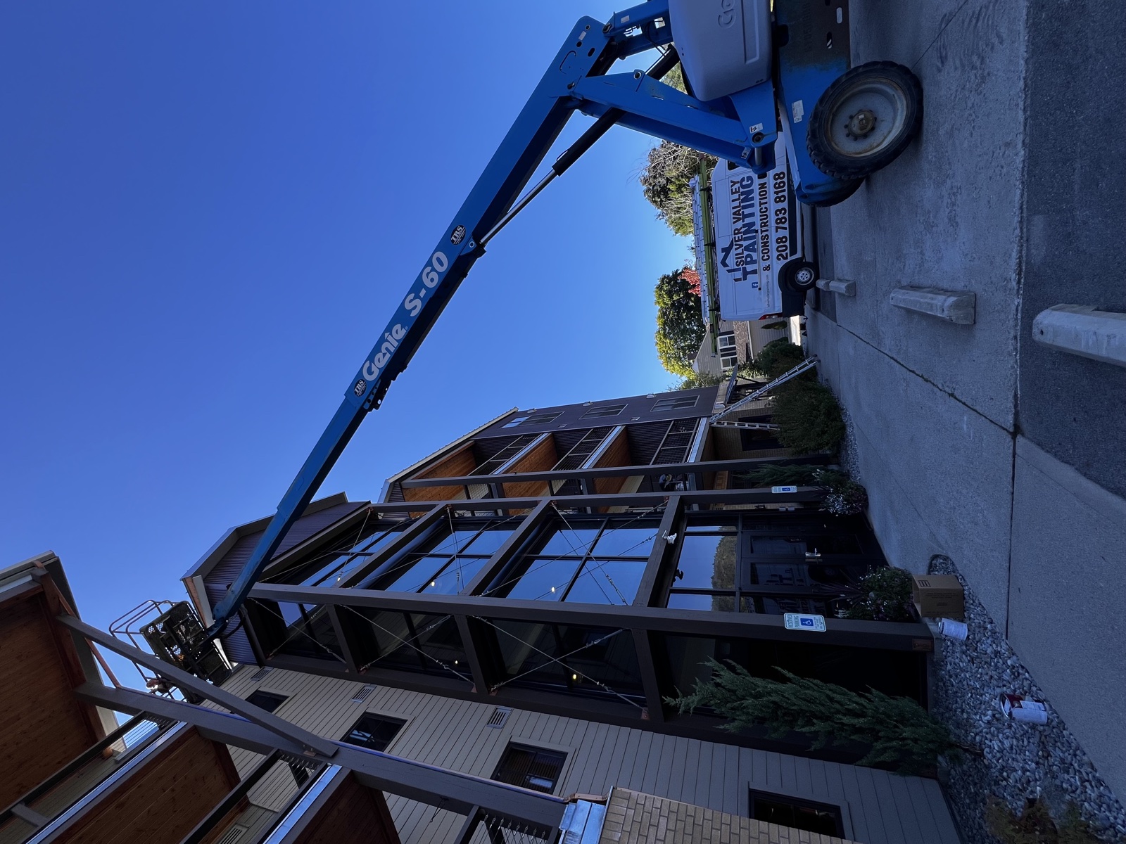 Modern dark-stained two-story home with large picture windows, mid-repaint with a blue articulating lift positioned in front and a Silver Valley work truck on the street