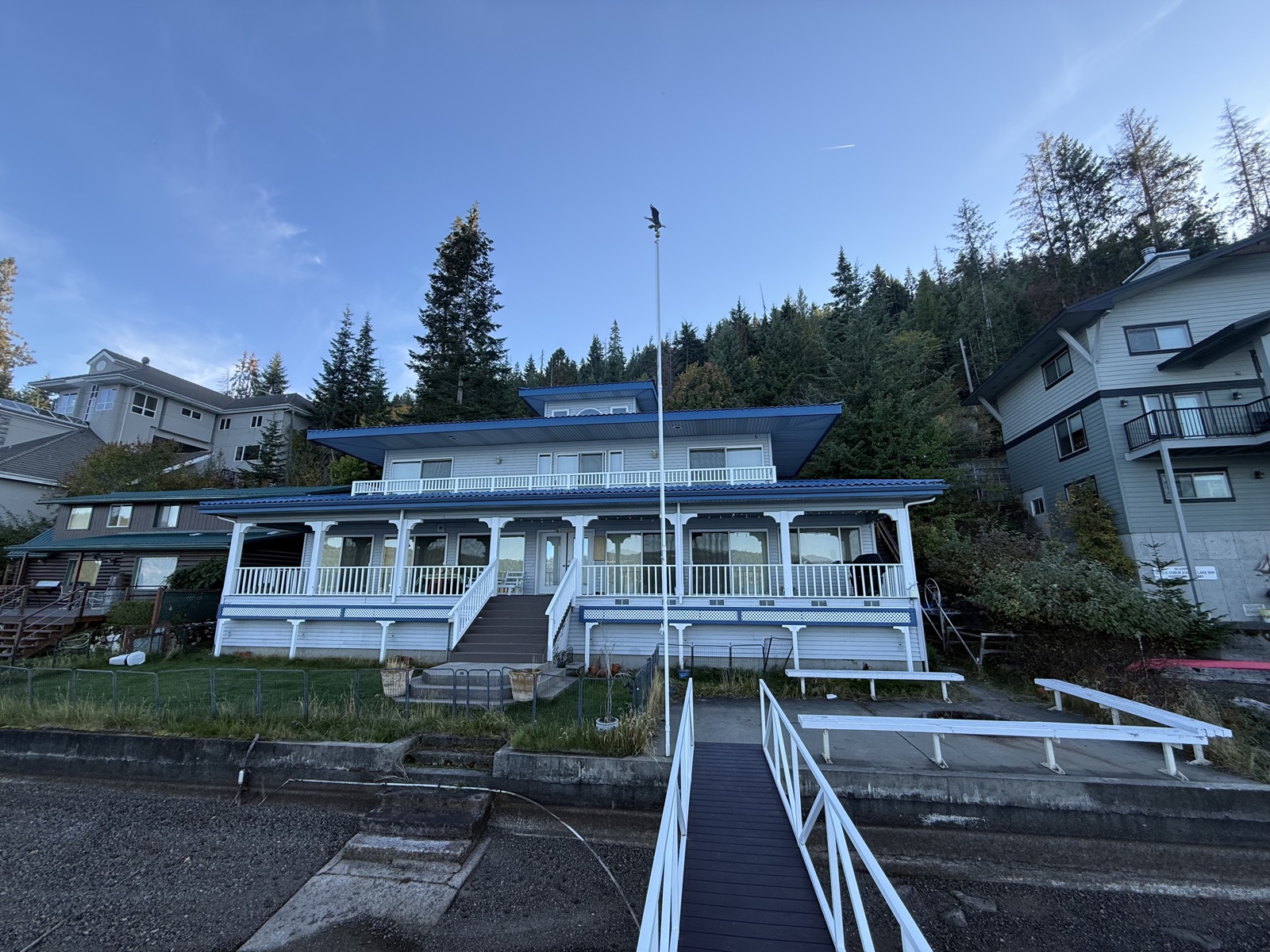 Two-story home with blue standing-seam roof, white siding, full wraparound covered porch with railings, set against a forested hillside