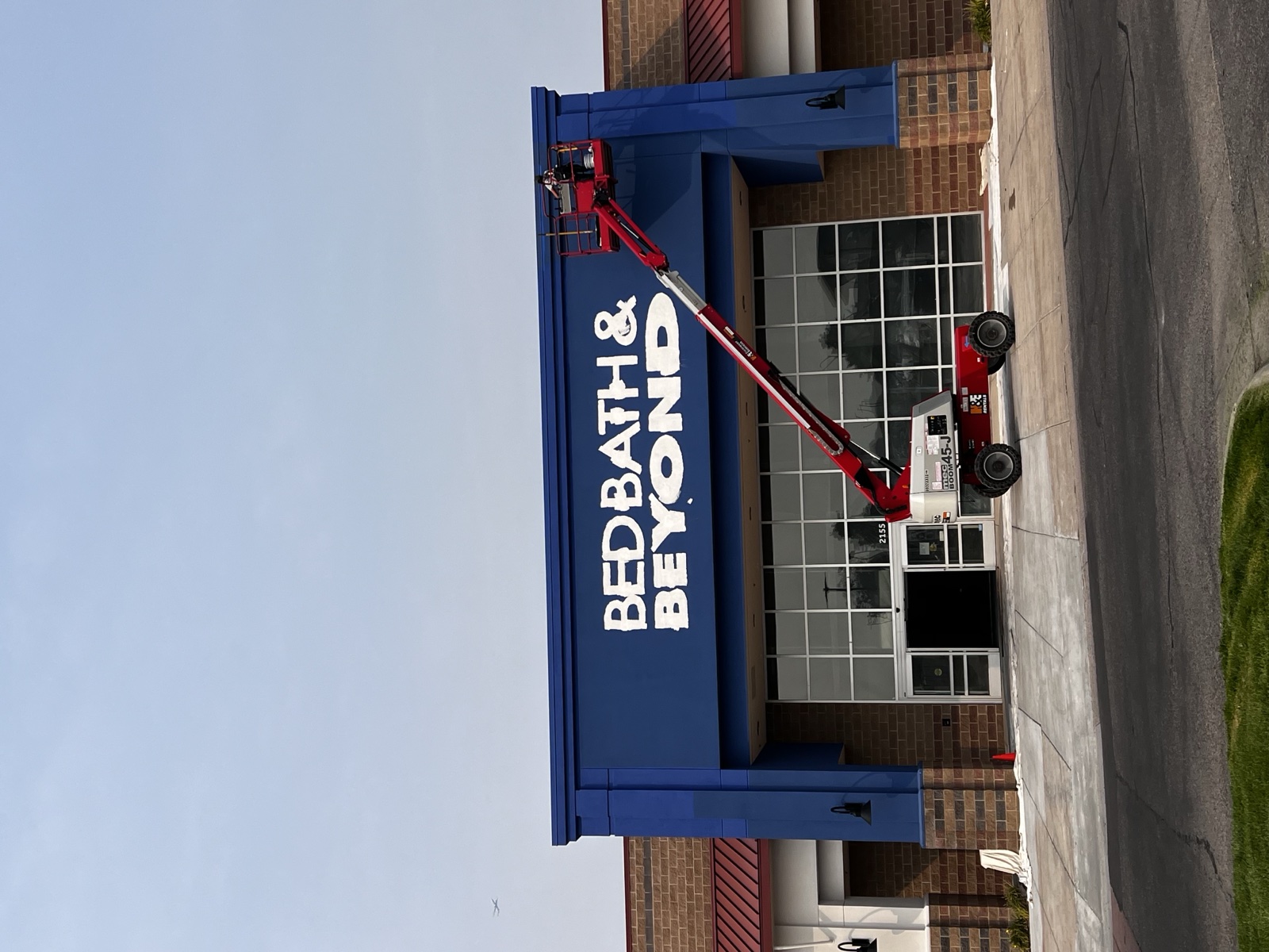 Closer view of a retail storefront mid-repaint with a Silver Valley painter on a red articulating lift coating the tall blue upper parapets above brick masonry