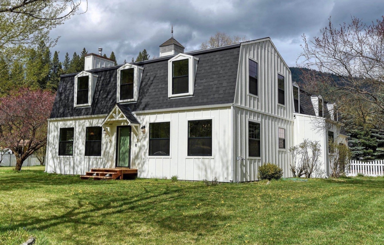 Two-story white farmhouse in Worley, Idaho with black gambrel-style roof, three dormers, and a small covered front entry against a dramatic cloudy sky