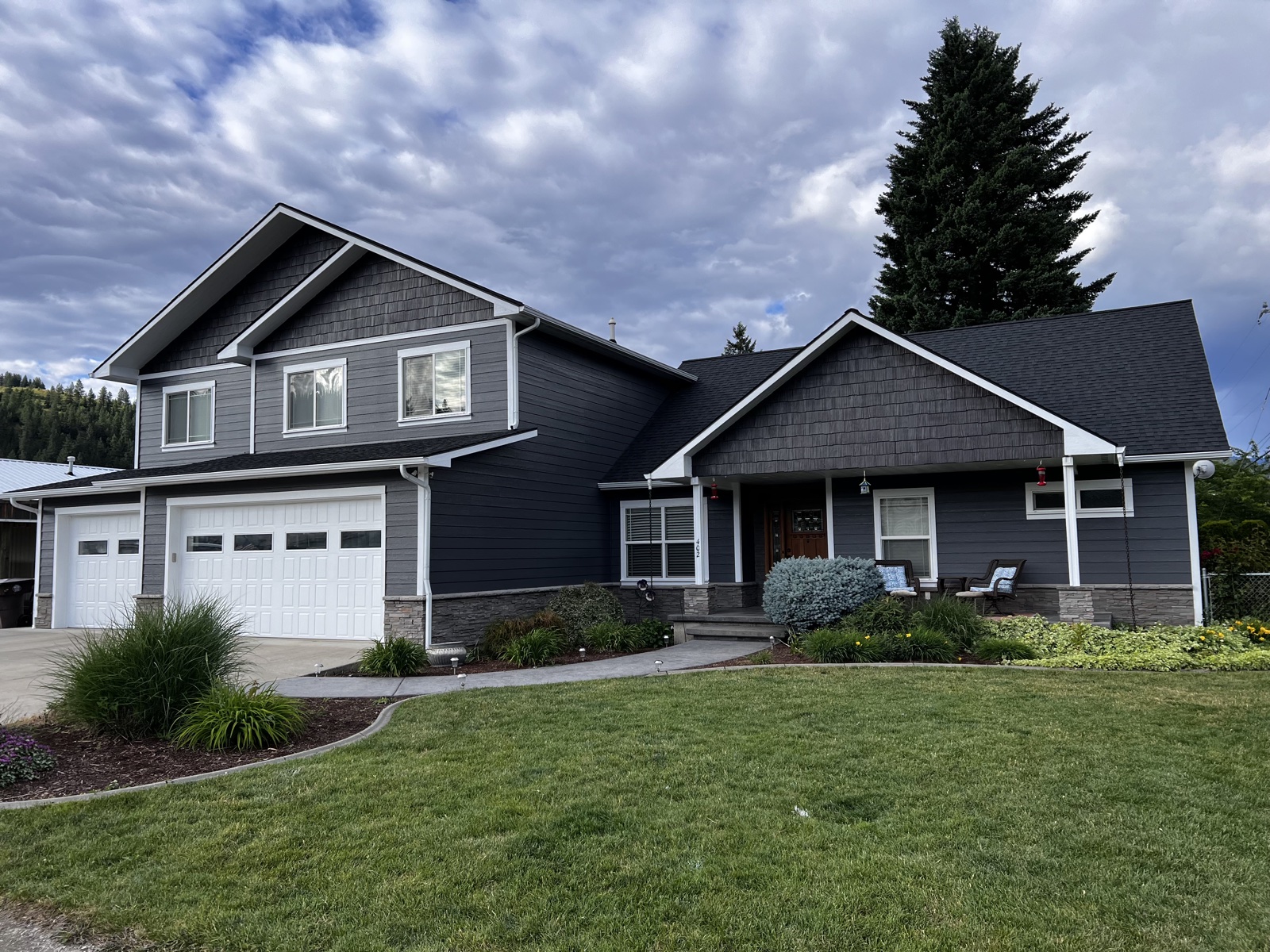 Two-story craftsman home in Hayden, Idaho with charcoal gray siding, attached two-car garage, stone porch detail, and curving paver walkway under a dramatic cloudy sky