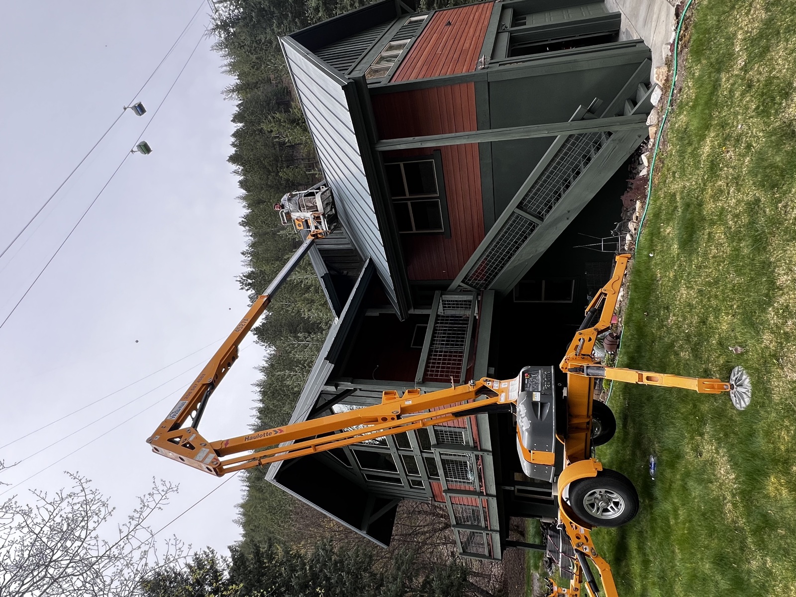 Modern dark gray home with cedar and red metal accents, mid-paint with a tall yellow articulating lift positioned for high-reach exterior coating