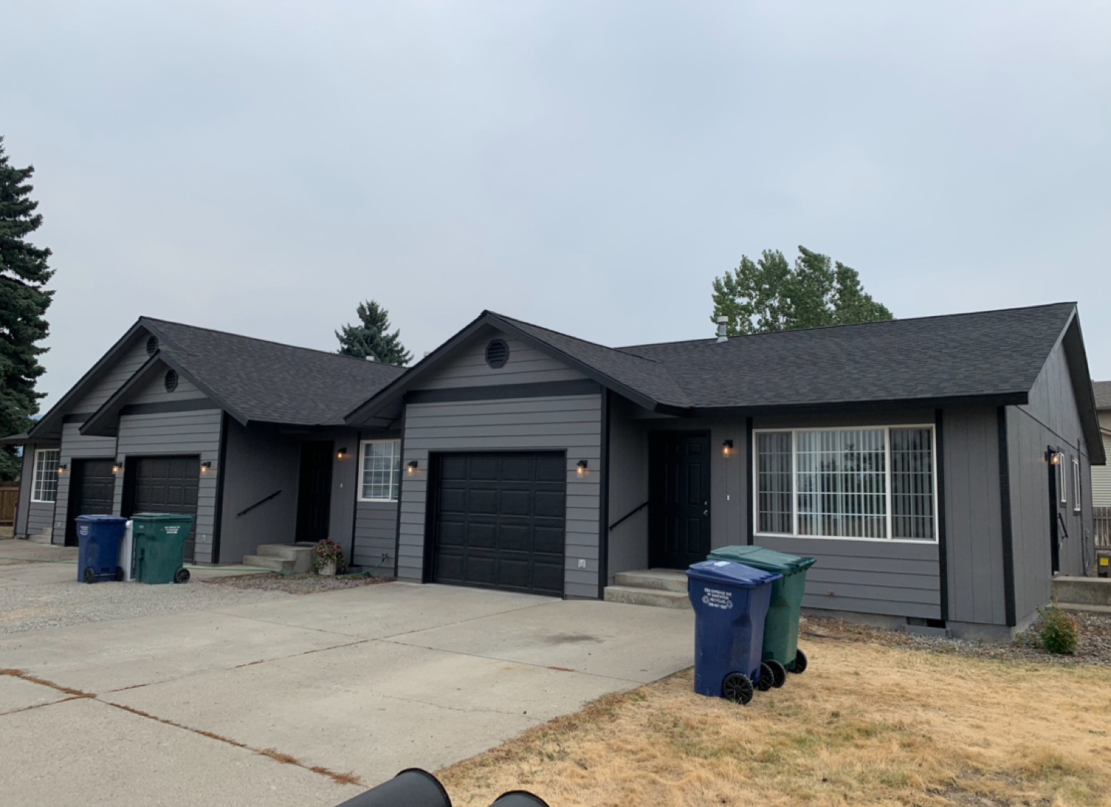 Single-story dark gray duplex in Post Falls, Idaho with crisp white window trim, double black garage doors, and trash bins on the driveway under a hazy sky
