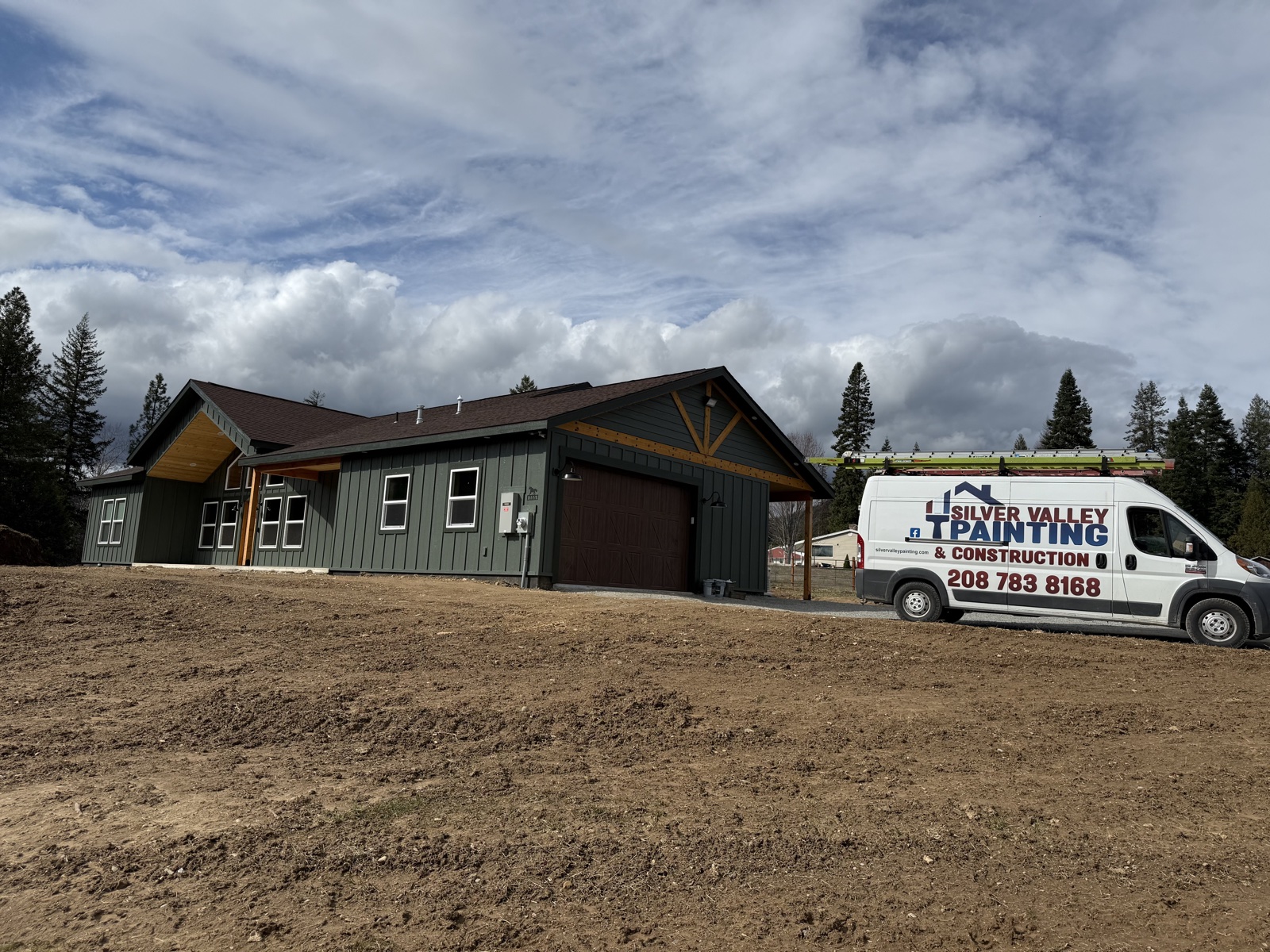 Single-story new-build home in Rathdrum, Idaho with dark green vertical siding, brown gabled roof, and a Silver Valley work van parked on the freshly graded site