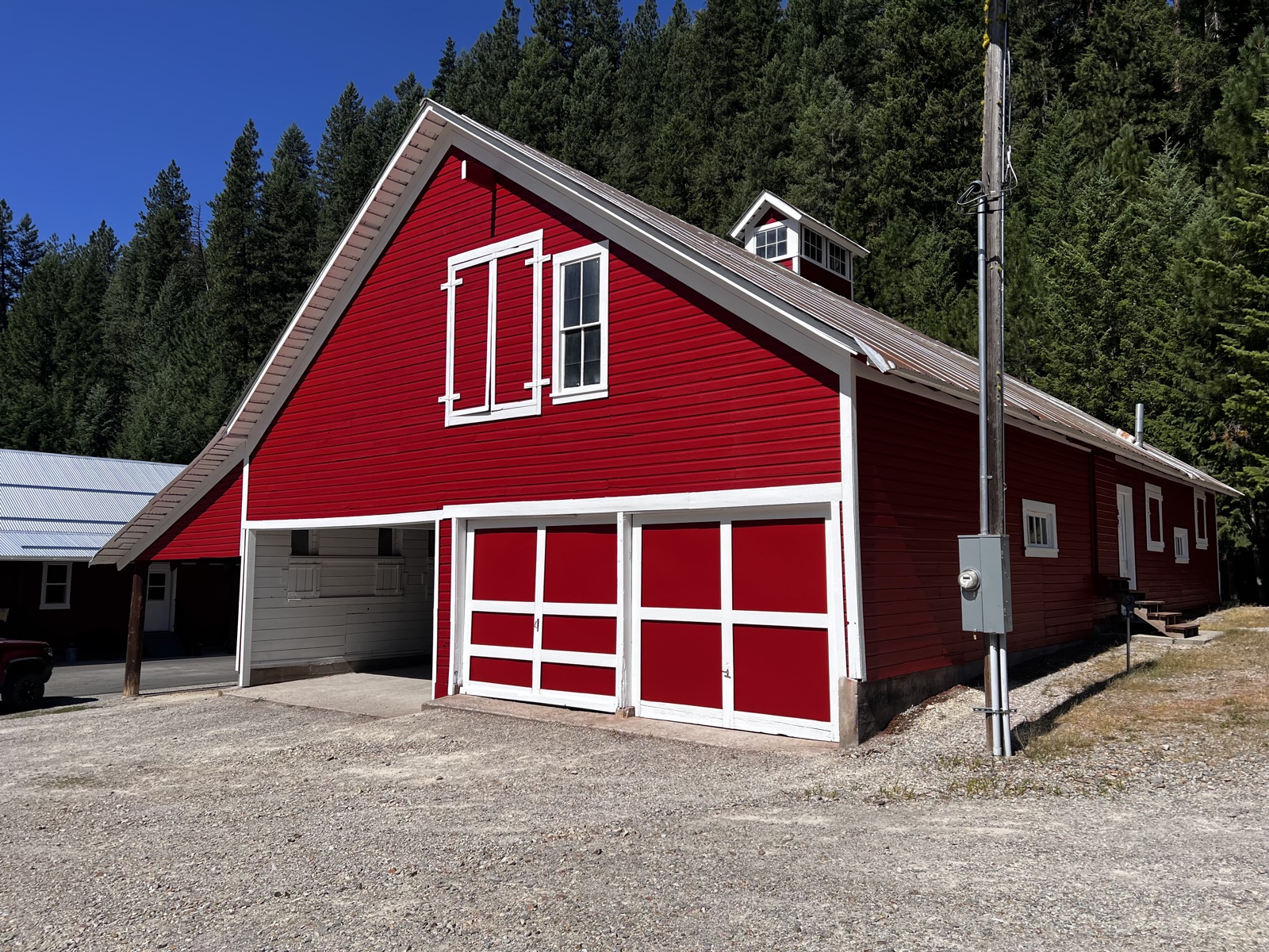 Classic red barn freshly painted with crisp white trim, double red garage doors, white-framed windows, and a forest of evergreens beyond