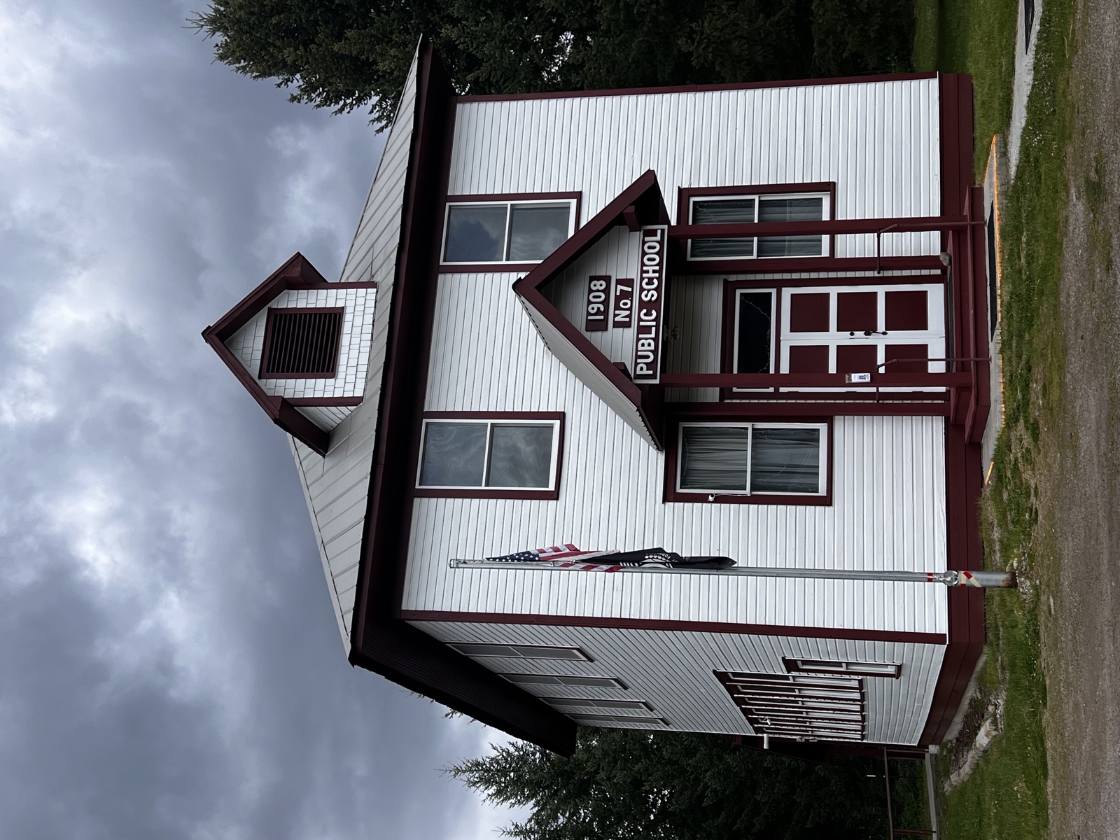 Front facade of the historic 1908 No. 7 Public Schoolhouse with fresh white siding, maroon-red trim and door, bell tower, and a flagpole out front