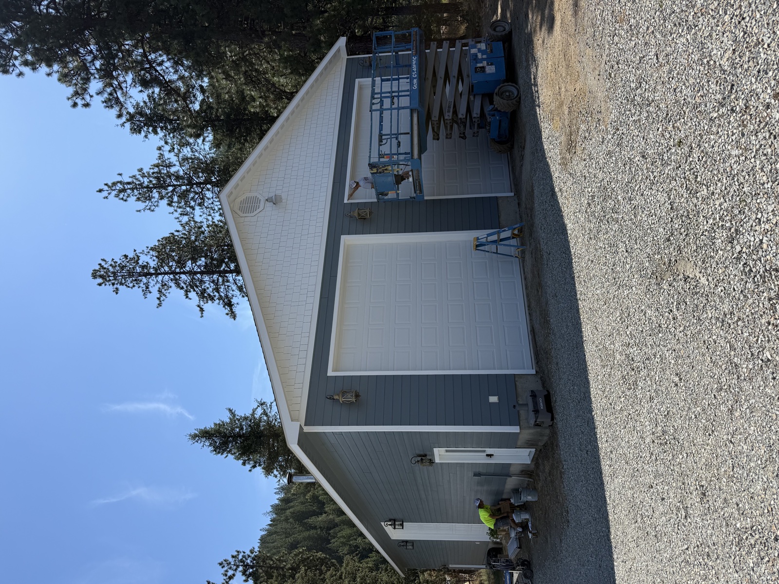 Long single-story commercial shop with white siding and dark gray accent panels, a scissor lift positioned for repainting overhead trim work, gravel lot in front