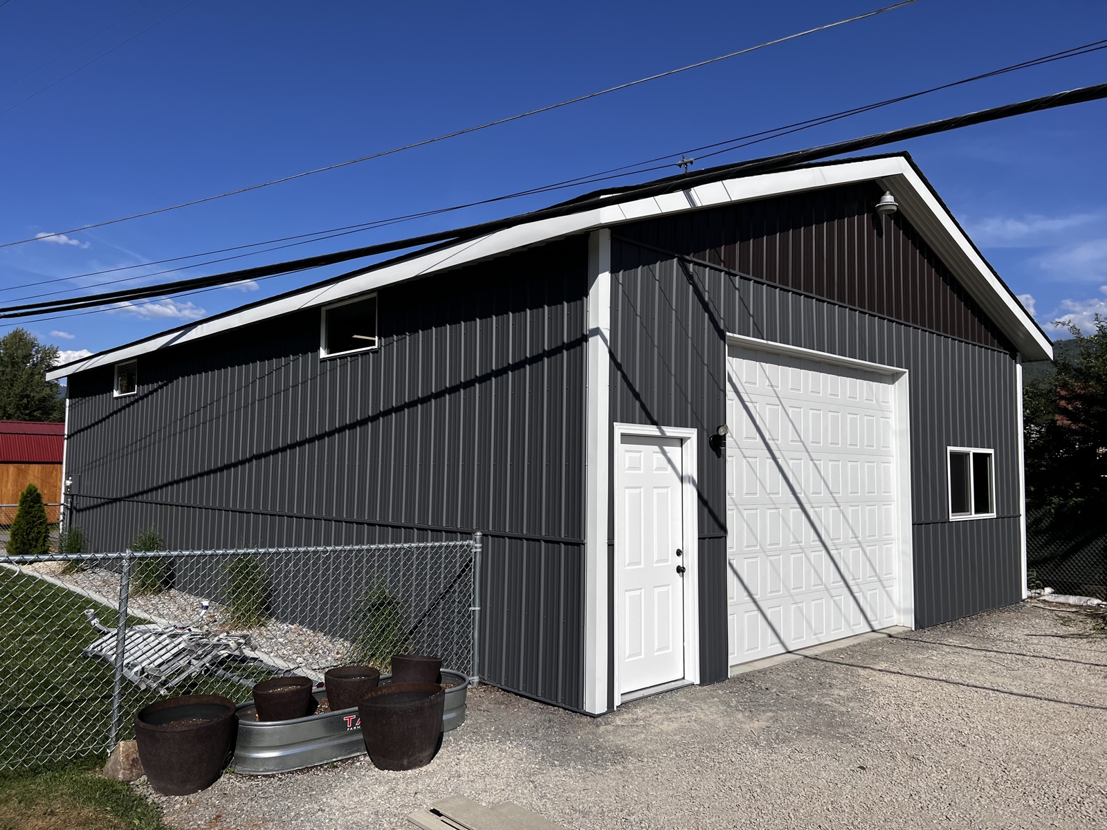 Dark gray metal shop building in Athol, Idaho with crisp white garage door, white trim, and galvanized stock-tank planters out front under a bright blue sky
