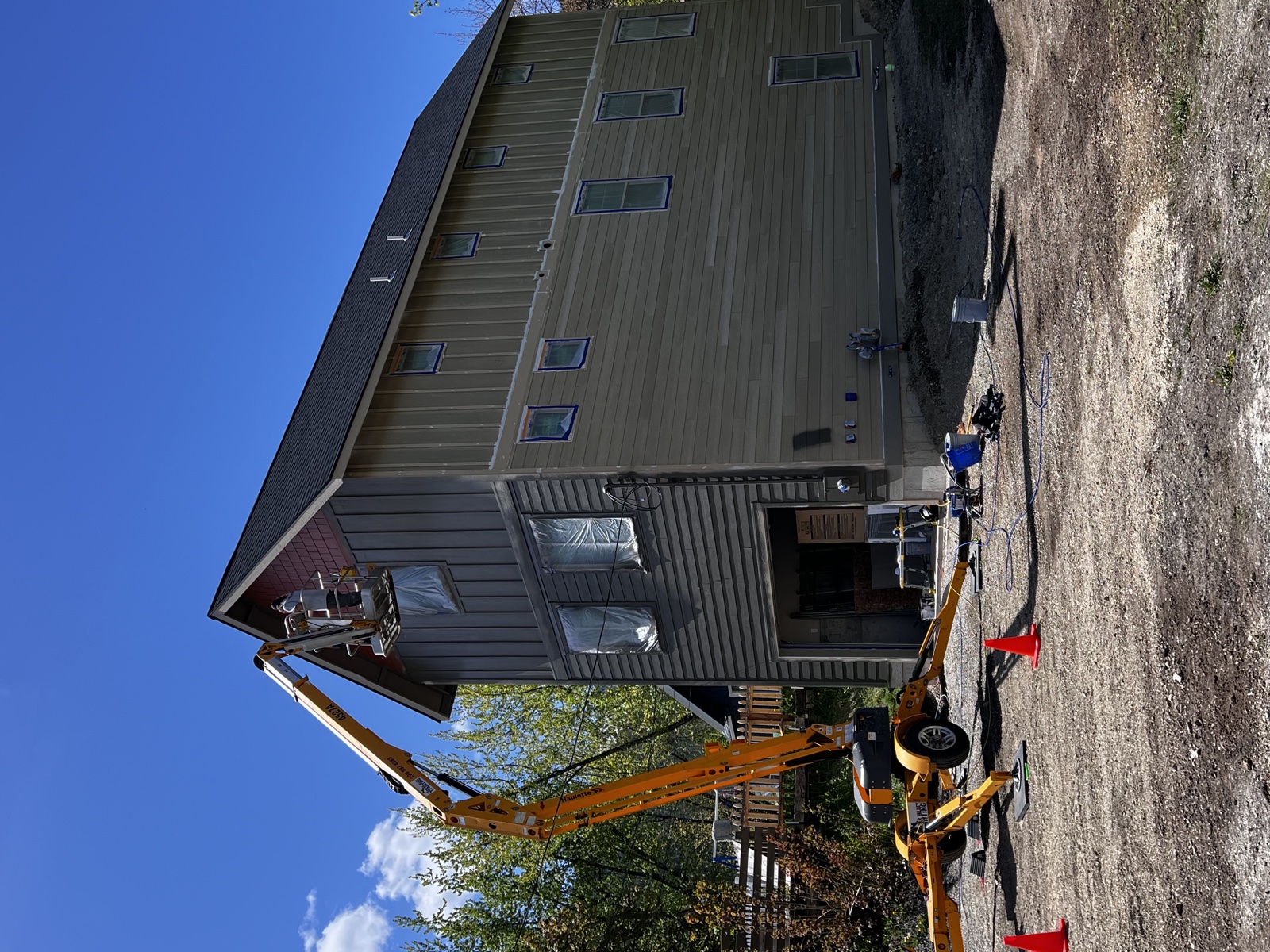 Modern dark-sided home with metal roof and tall windows mid-exterior repaint, with an orange articulating lift staged on the gravel driveway