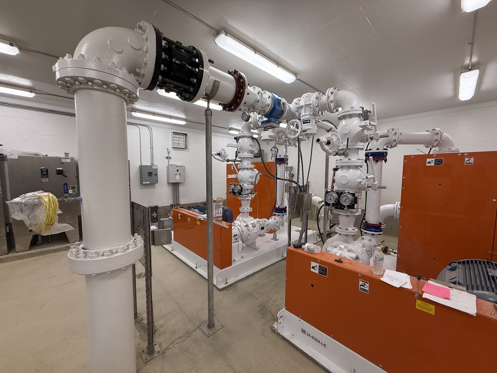 Municipal pump station interior with newly epoxy-coated white process piping, blue valves, and orange industrial equipment housings on a concrete floor