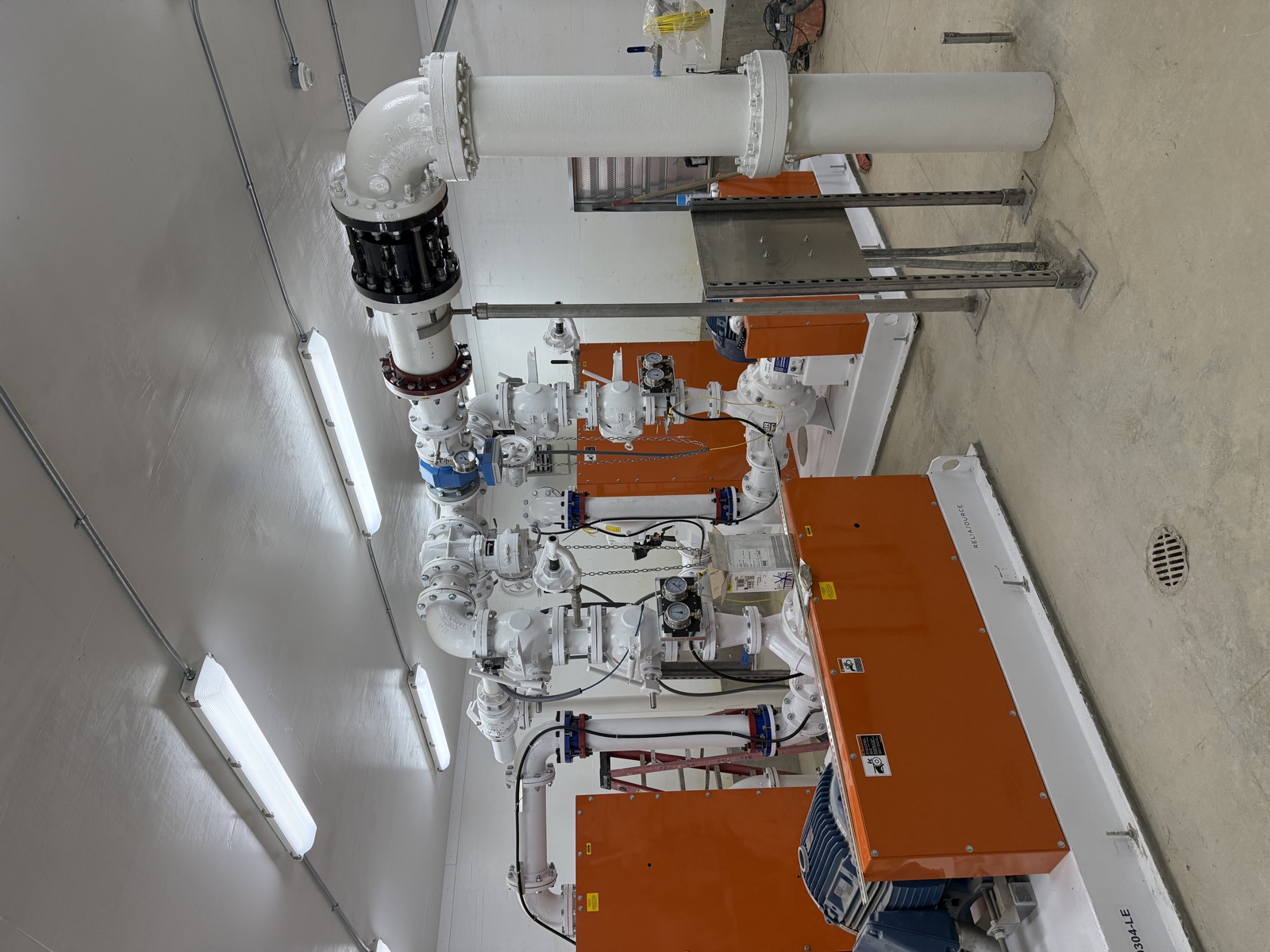 Top-down view of a wastewater lift station with epoxy-coated white piping and orange-painted pump housings beneath fluorescent lighting