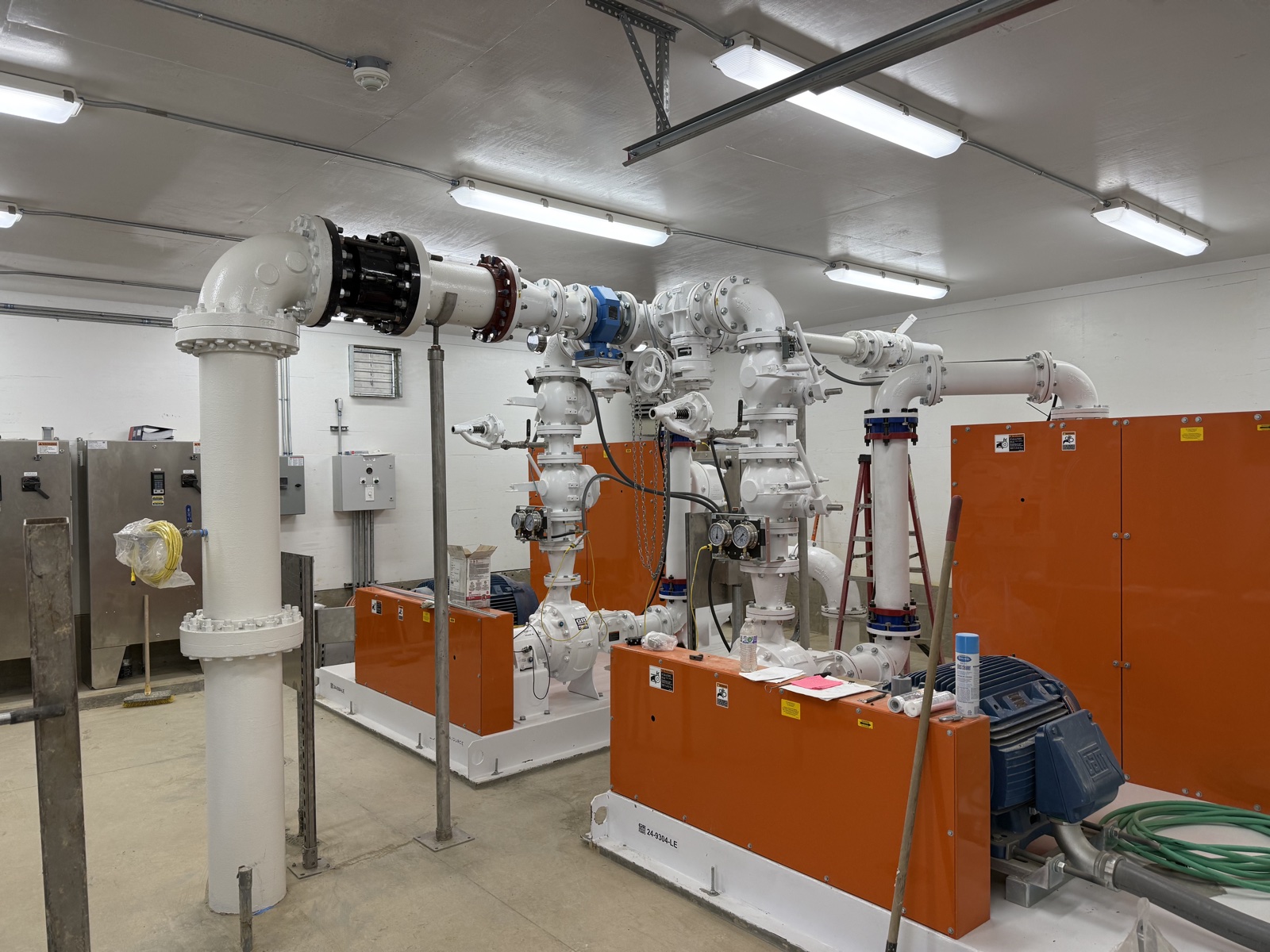 Epoxy-coated white pipe manifold with blue valves and orange pump-enclosure panels in a wastewater treatment facility service room