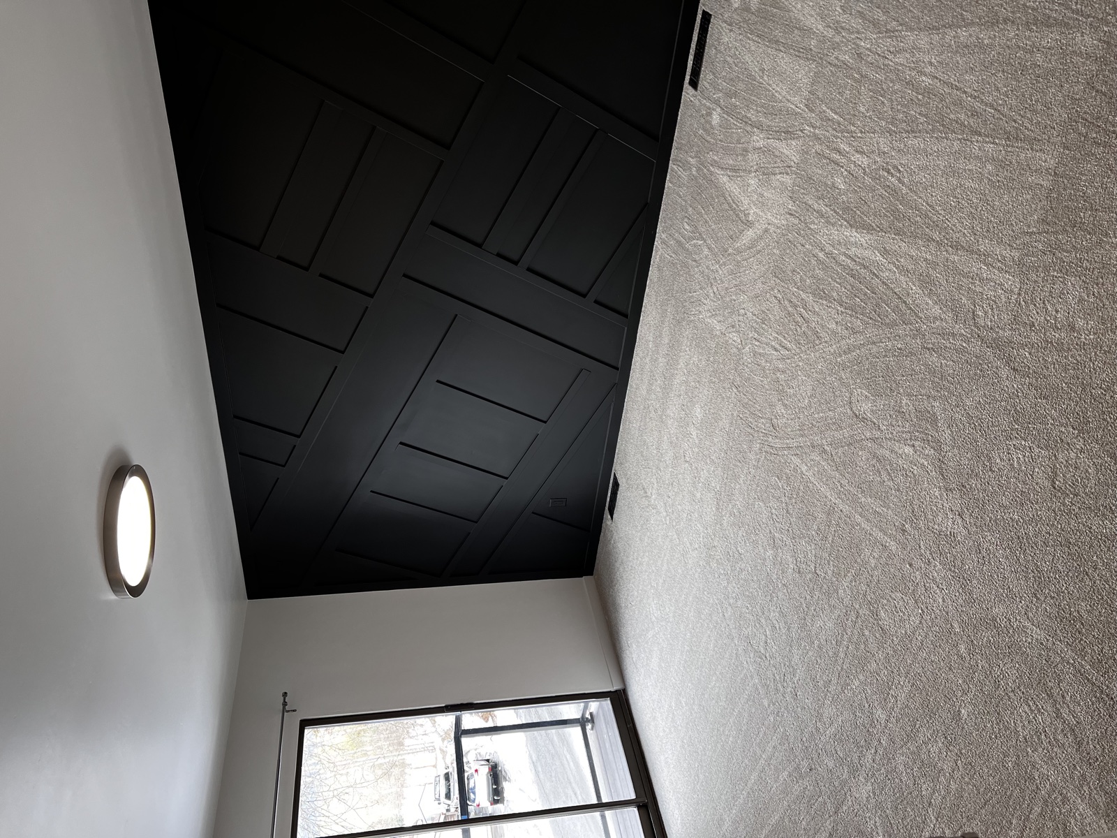 Two-story foyer featuring a dramatic black geometric grid-pattern accent wall paired with a textured silver-gray plaster wall and modern round flush-mount light