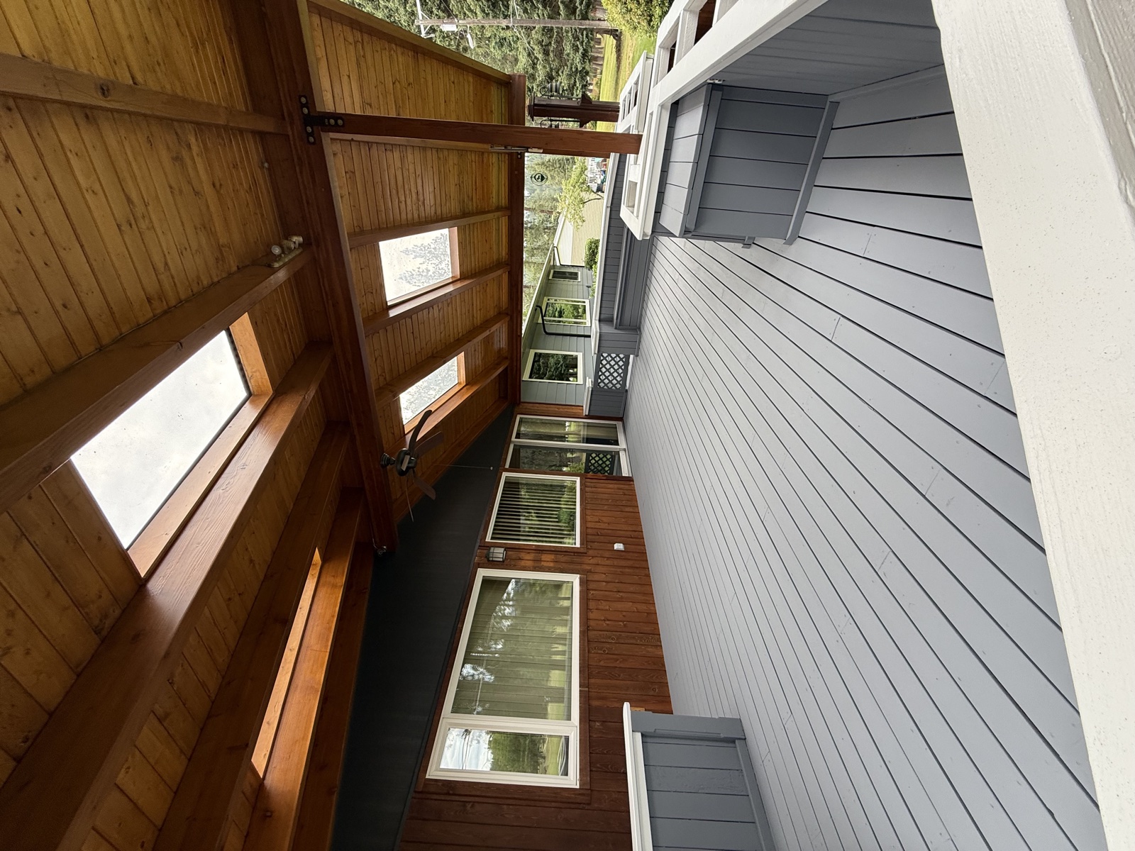 Custom covered outdoor porch with warm cedar tongue-and-groove ceiling, gray painted siding, and a row of large picture windows looking out to the yard