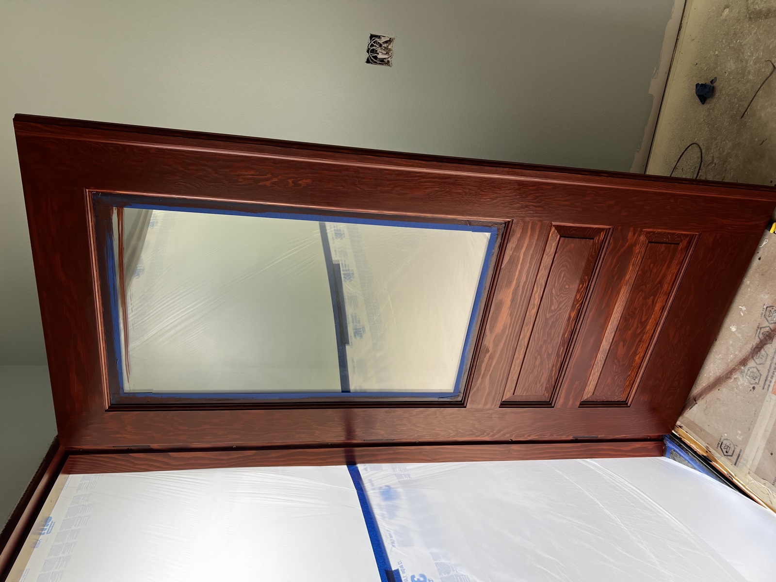 Solid hardwood entry door freshly stained in a rich mahogany tone with raised panels and a frosted glass window light, taped off for the finish coat