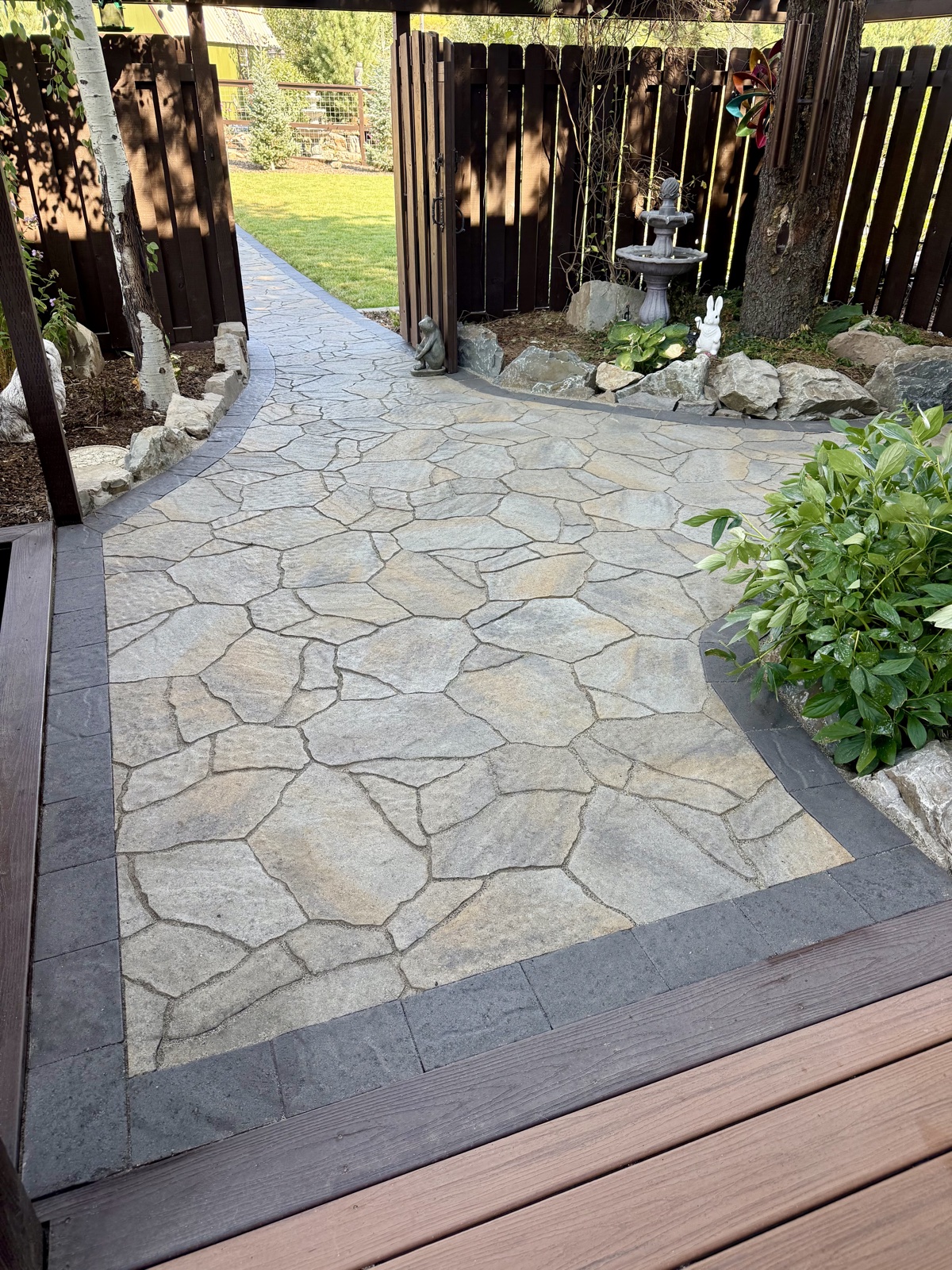 Completed irregular flagstone patio with charcoal stone border, garden fountain, surrounding shrubs, and a dark wood privacy fence backdrop