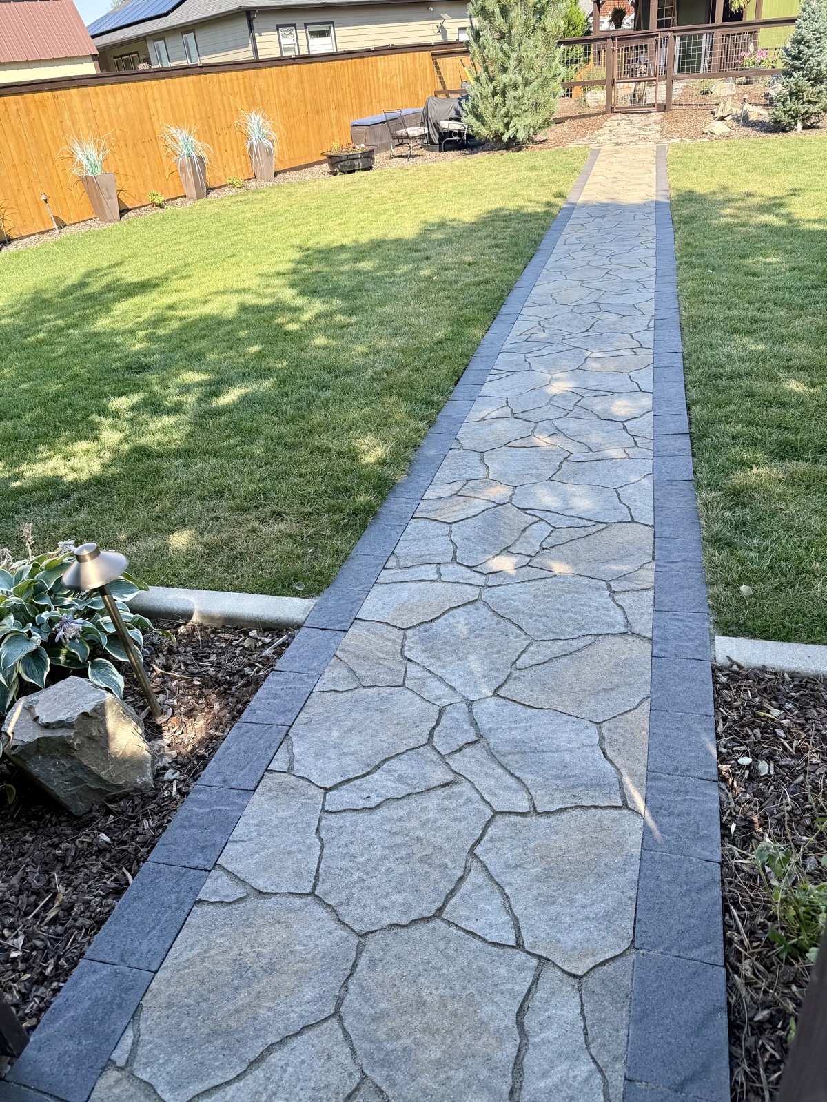 Long flagstone walkway with a dark charcoal stone border running through a manicured backyard toward a deck, with privacy fence and lawn beyond