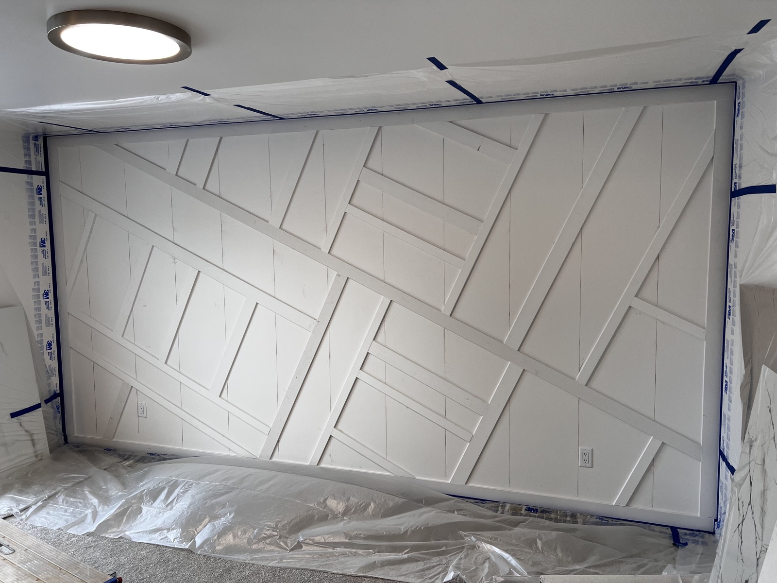 Custom geometric criss-cross trim accent wall painted crisp white, taped off and floor-protected during finish coating, with a flush dome light above