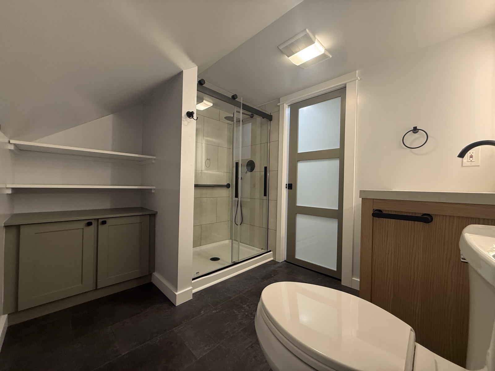 Compact bathroom remodel with white walls, a sage green shaker vanity, glass shower with subway tile, white wainscot accent, and dark slate tile floor
