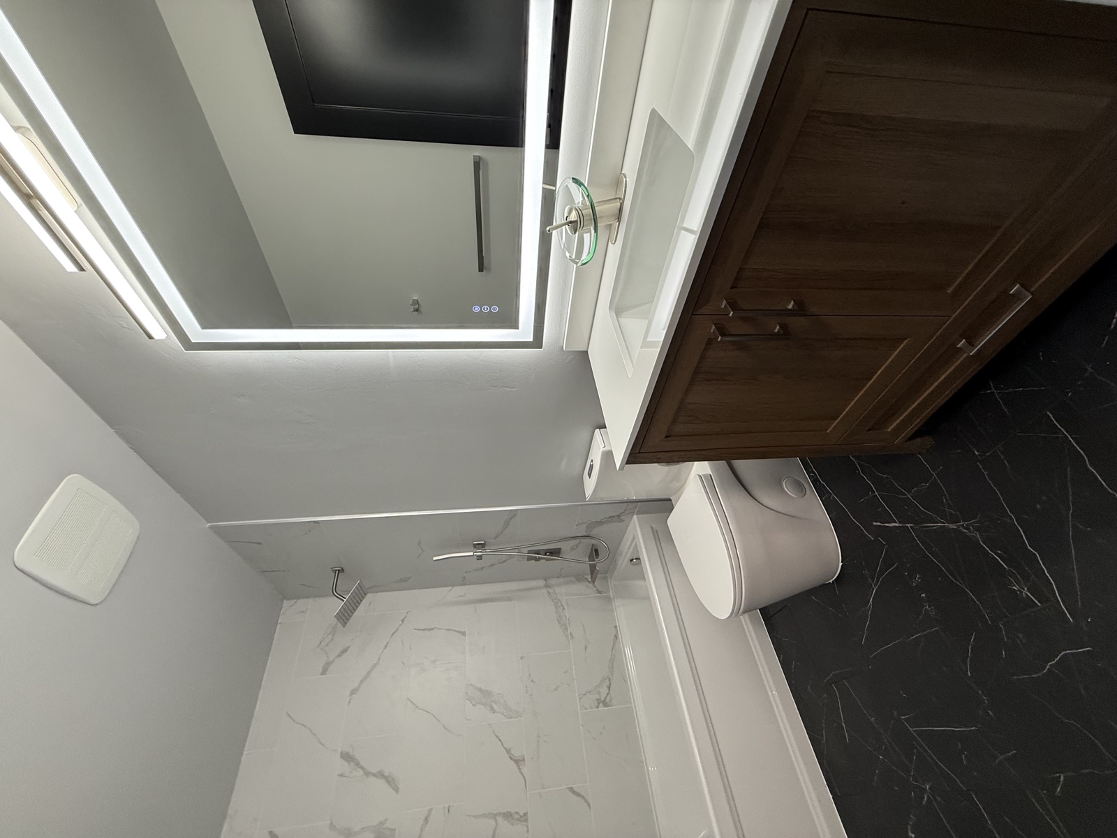Compact powder room with dramatic black marbled tile feature wall and matching floor, a walnut vanity, illuminated LED mirror, and wall-mounted sink