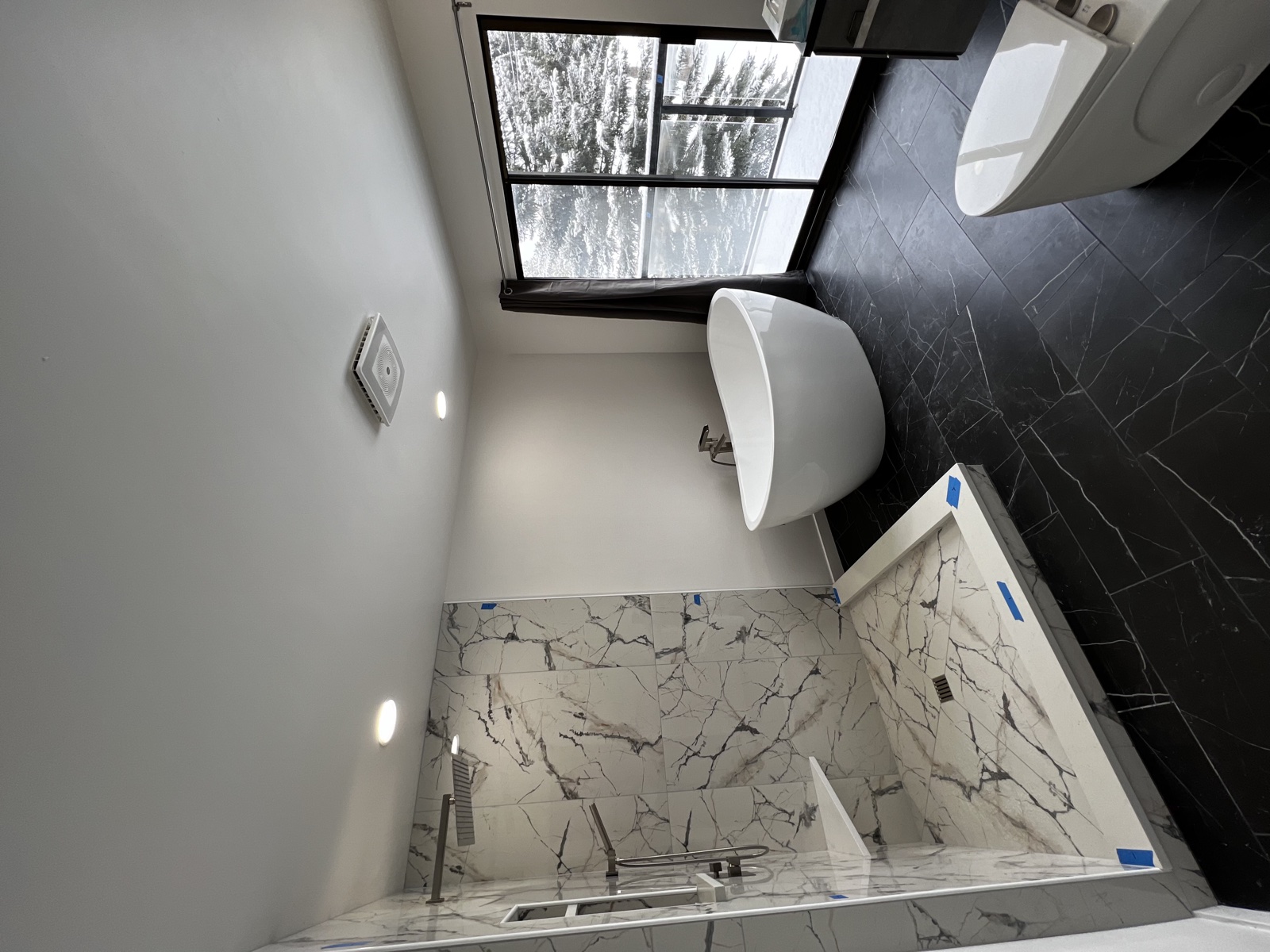 Spa-style bathroom under finish work with marble slab tub surround, modern wall-mounted tub filler, and a tall bay window, blue painter's tape still in place