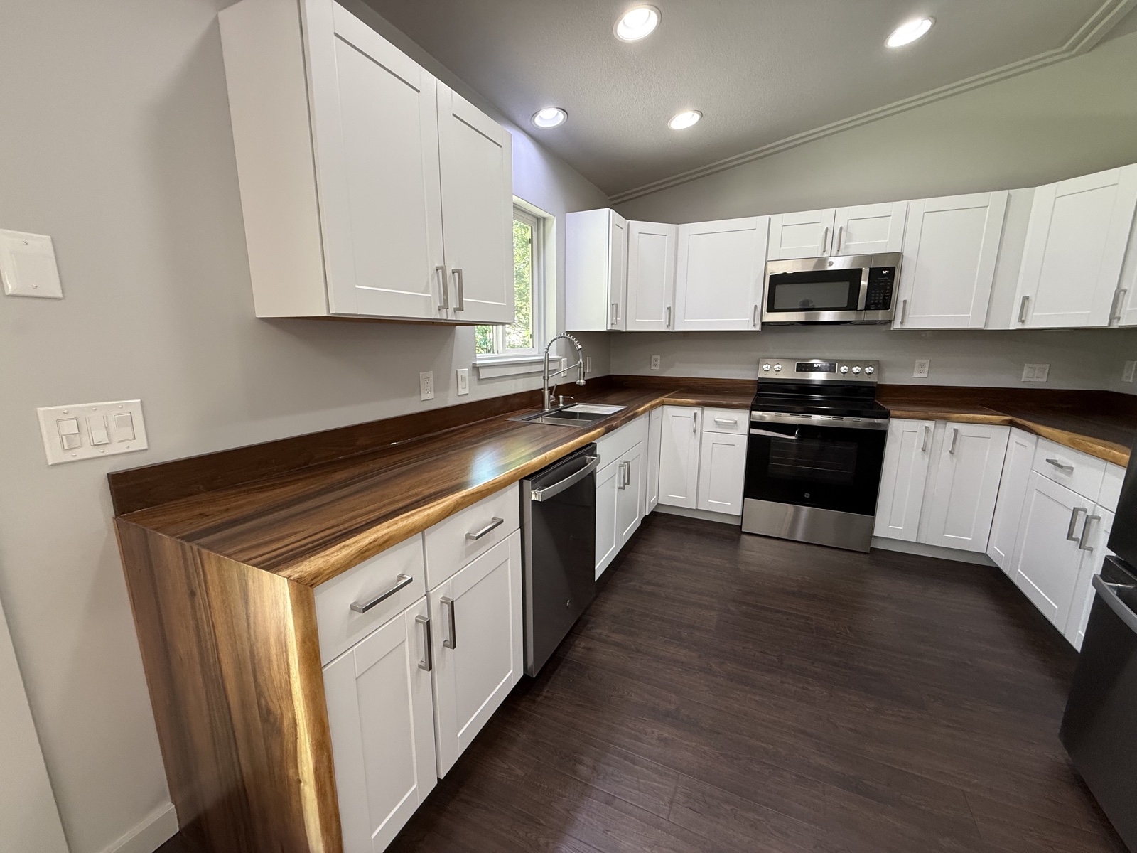 Open-concept kitchen and great room with white shaker cabinets, a live-edge wood counter, stainless appliances, and a modern circular ringed pendant over a dark wood floor
