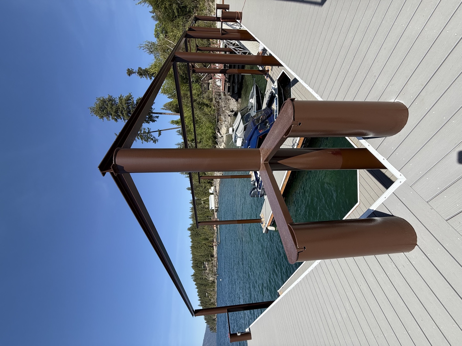 Refinished lakefront wood dock with chocolate brown wrap on each support piling, smooth gray plank decking, and forested lake shoreline beyond