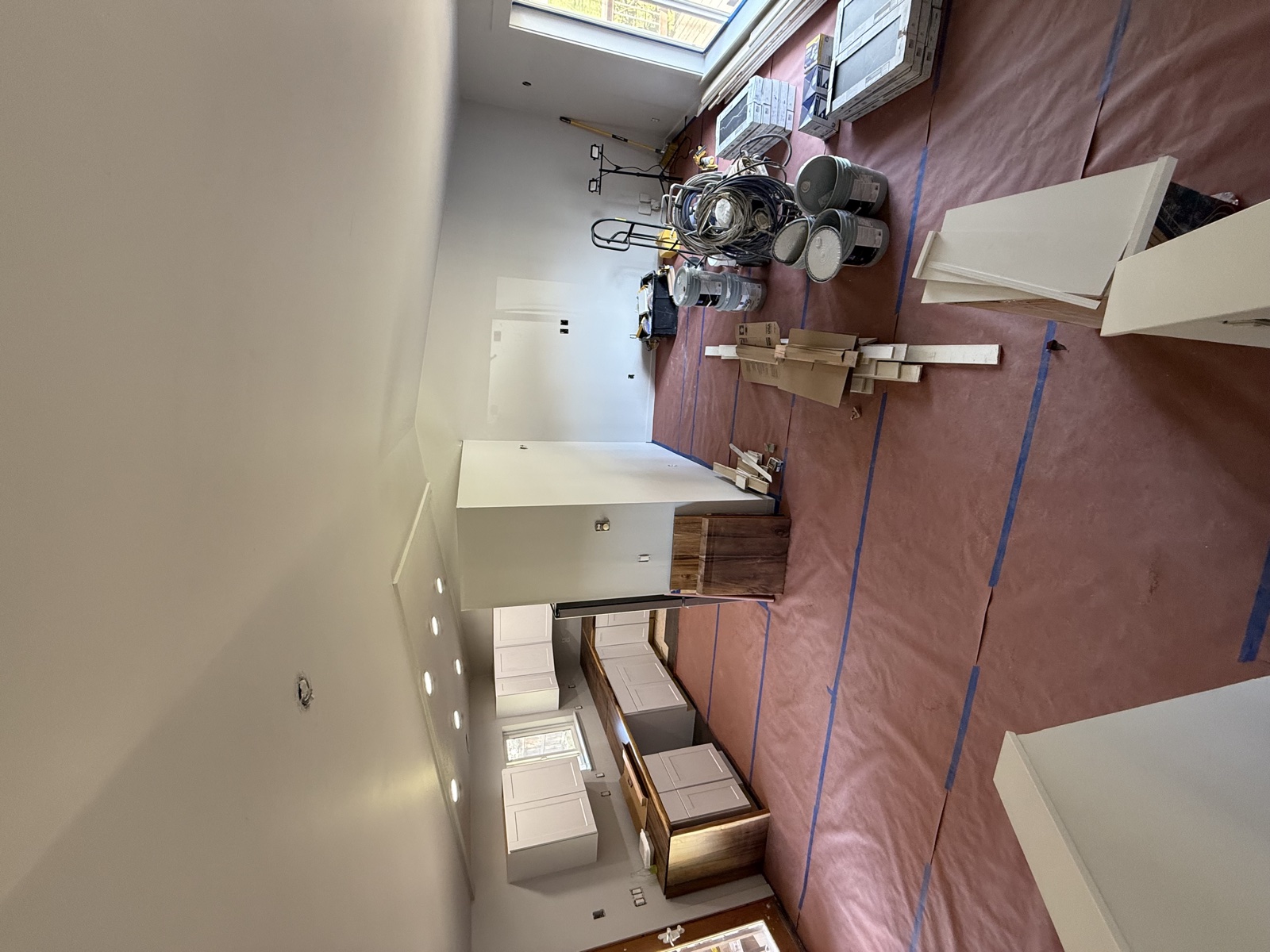 Kitchen mid-remodel with finished white shaker cabinets installed against pink insulation paper, drawer fronts stacked nearby, and paint cans and floor protection in place