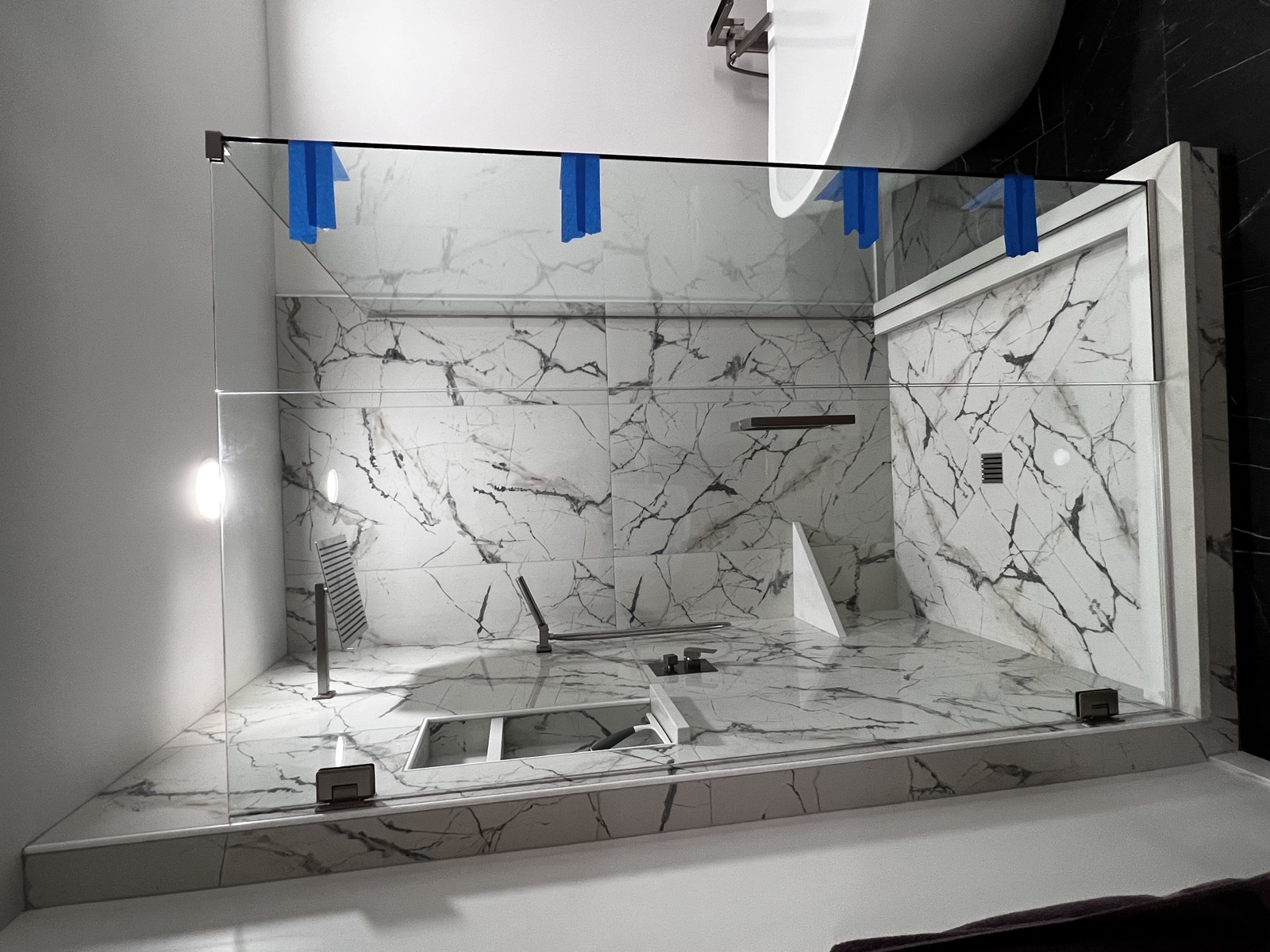 Detail of a white marble tub surround with built-in handheld sprayer and tub filler, blue painter's tape still in place along the upper edge