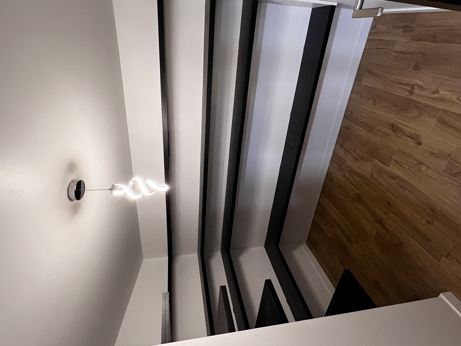 Modern stairwell remodel detail with twisting glass-tube pendant sconce, black steel stair rails, and bright white walls over warm wood flooring