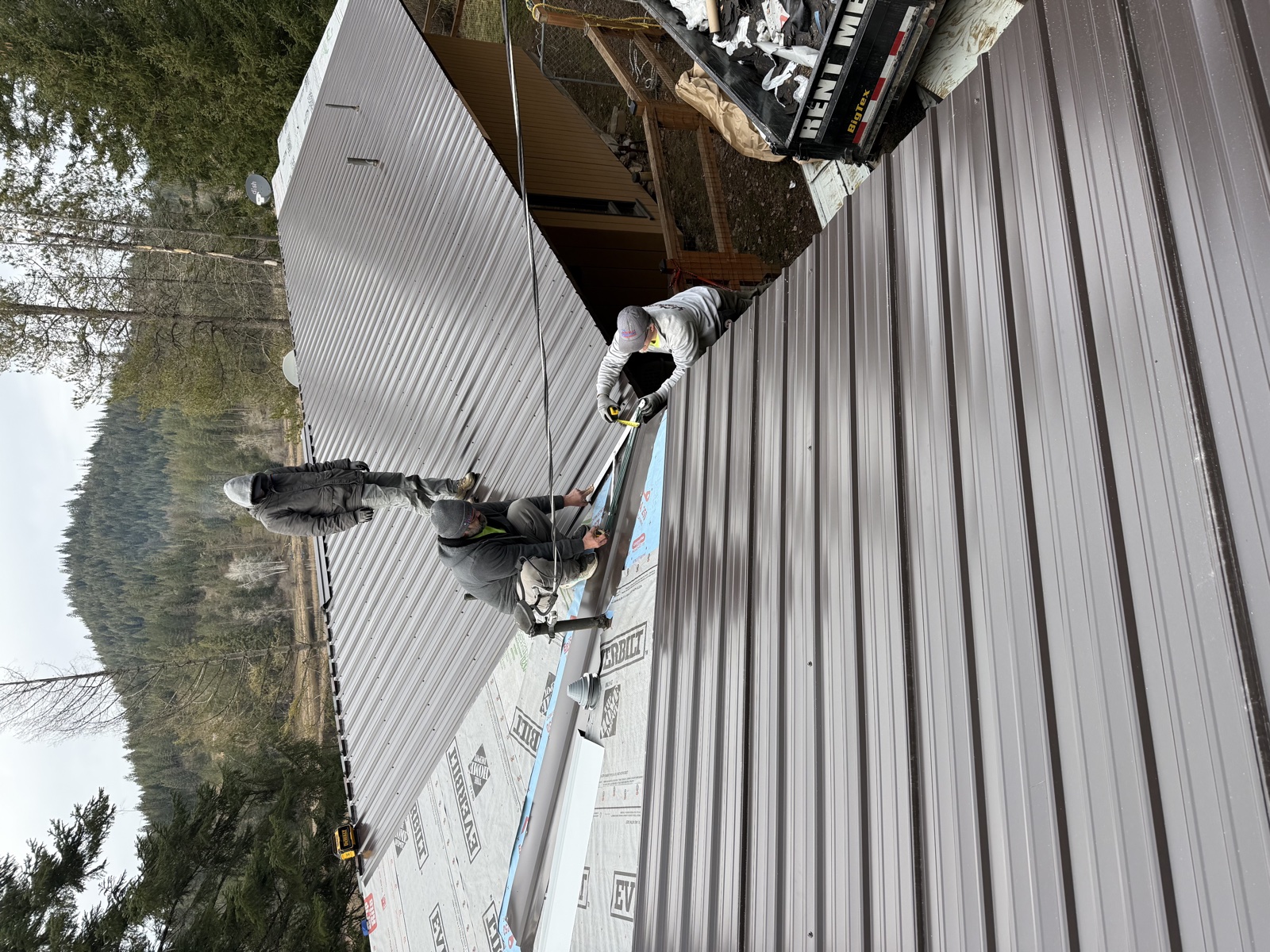Two Silver Valley crew members installing a gray standing-seam metal roof on a Pinehurst, Idaho home with construction wrap visible on the gables